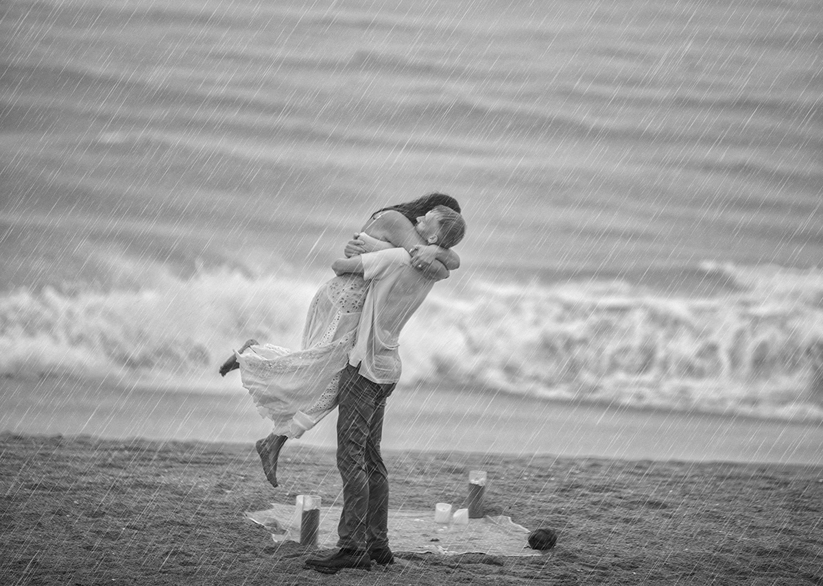 Couple embracing during a rain-soaked beach proposal, with waves crashing behind them.