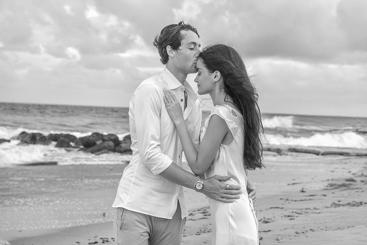 Couple embracing on a Florida beach after a marriage proposal, groom kissing fiancée’s forehead as waves crash behind them.