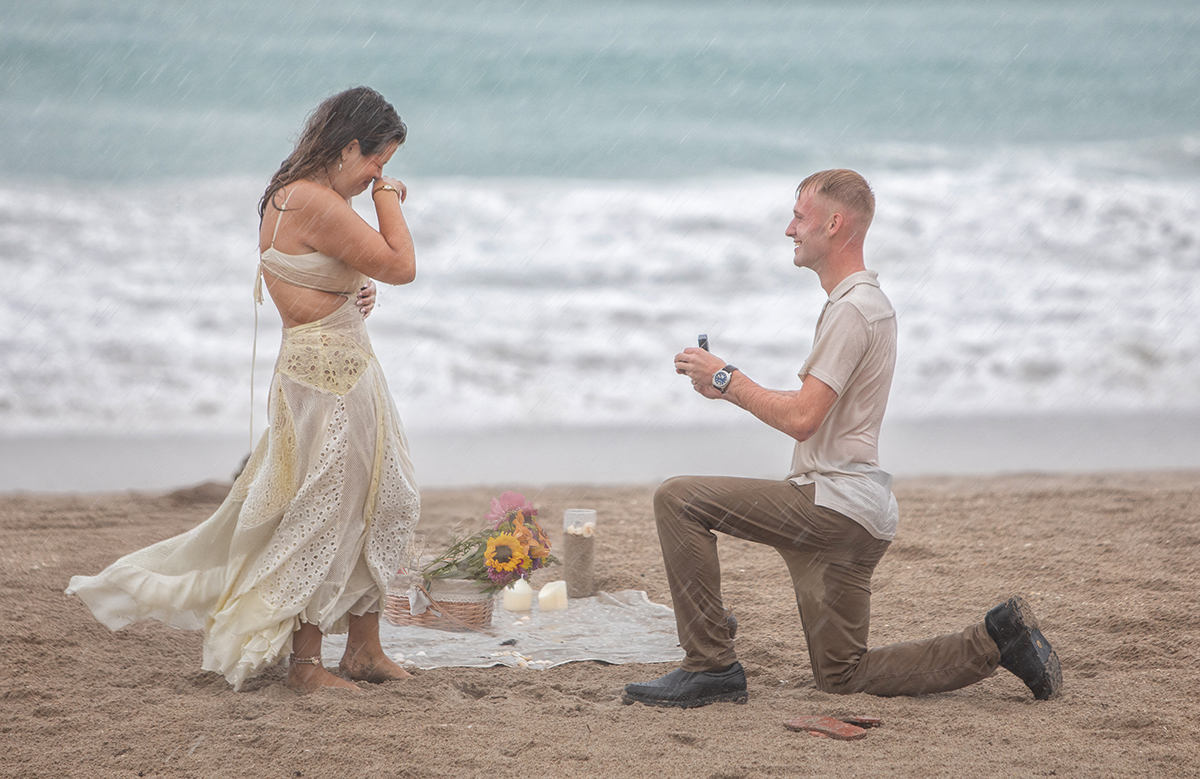 A Florida proposal photographer captures an emotional beach engagement during a surprise proposal in the rain, with the groom kneeling in wet sand as the bride-to-be covers her face in tears of joy.