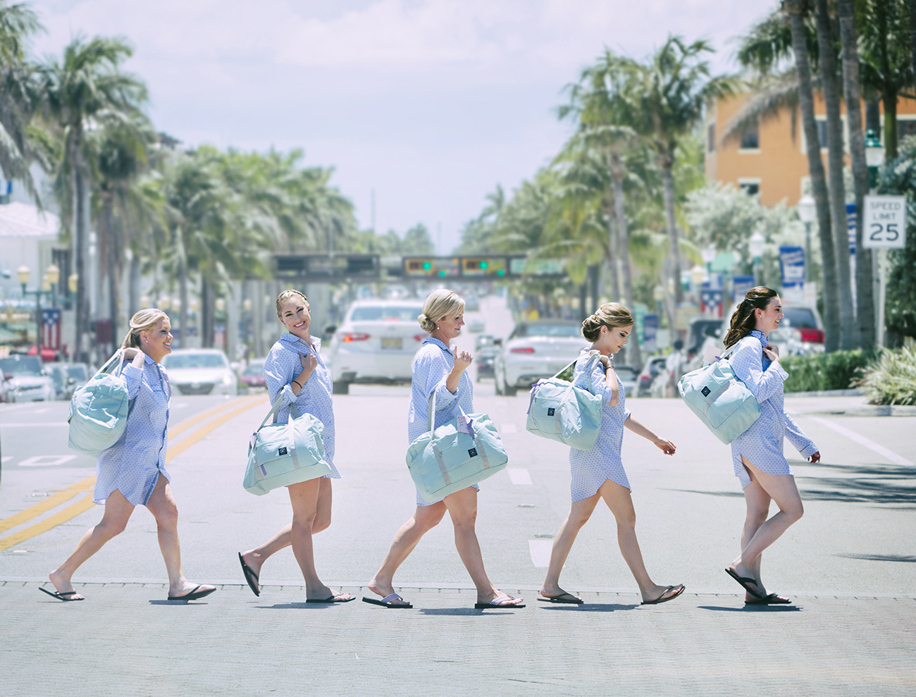 Bridesmaids cross Ocean Boulevard in Delray Beach, walking from the Opal Grand Resort toward the beach. They wear matching light blue shirts and carry mint duffel bags, smiling under the South Florida sun with palm trees and traffic in the background.