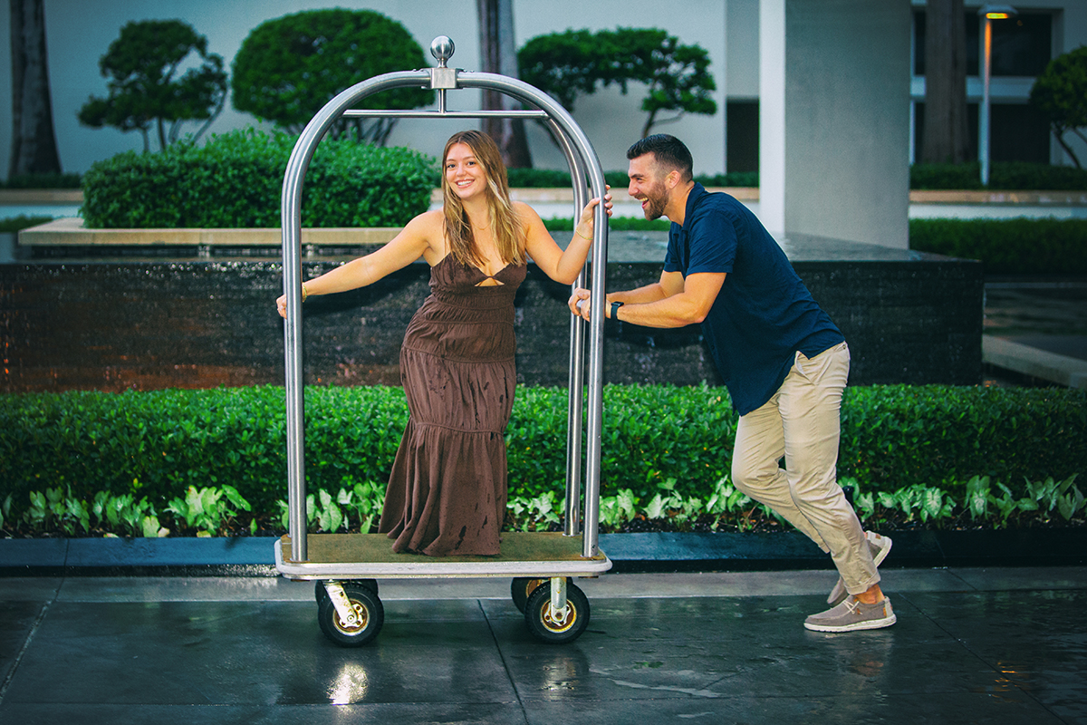 A newly engaged couple laughing and playing on a hotel luggage cart after a rainy South Florida marriage proposal, with the bride-to-be riding while her fiancé playfully pushes.