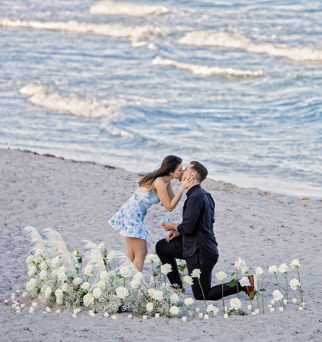 Couple kissing on the beach after a romantic proposal surrounded by white roses at South Beach Park in Boca Raton, captured by Florida marriage proposal photographer Jeff Kolodny.