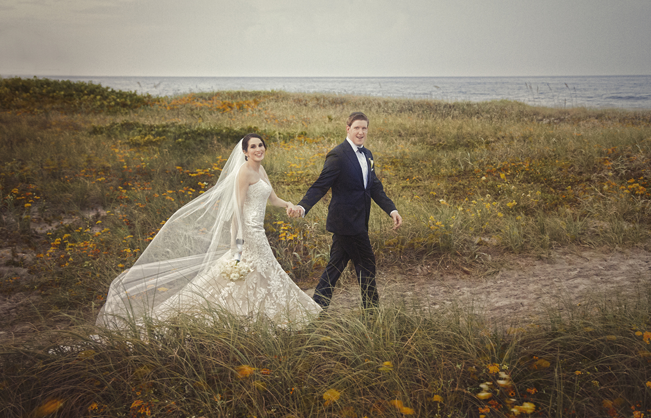 Bride and groom walking hand-in-hand along a wildflower-lined beach path near the Opal Grand in South Florida, with the bride’s veil flowing in the ocean breeze.