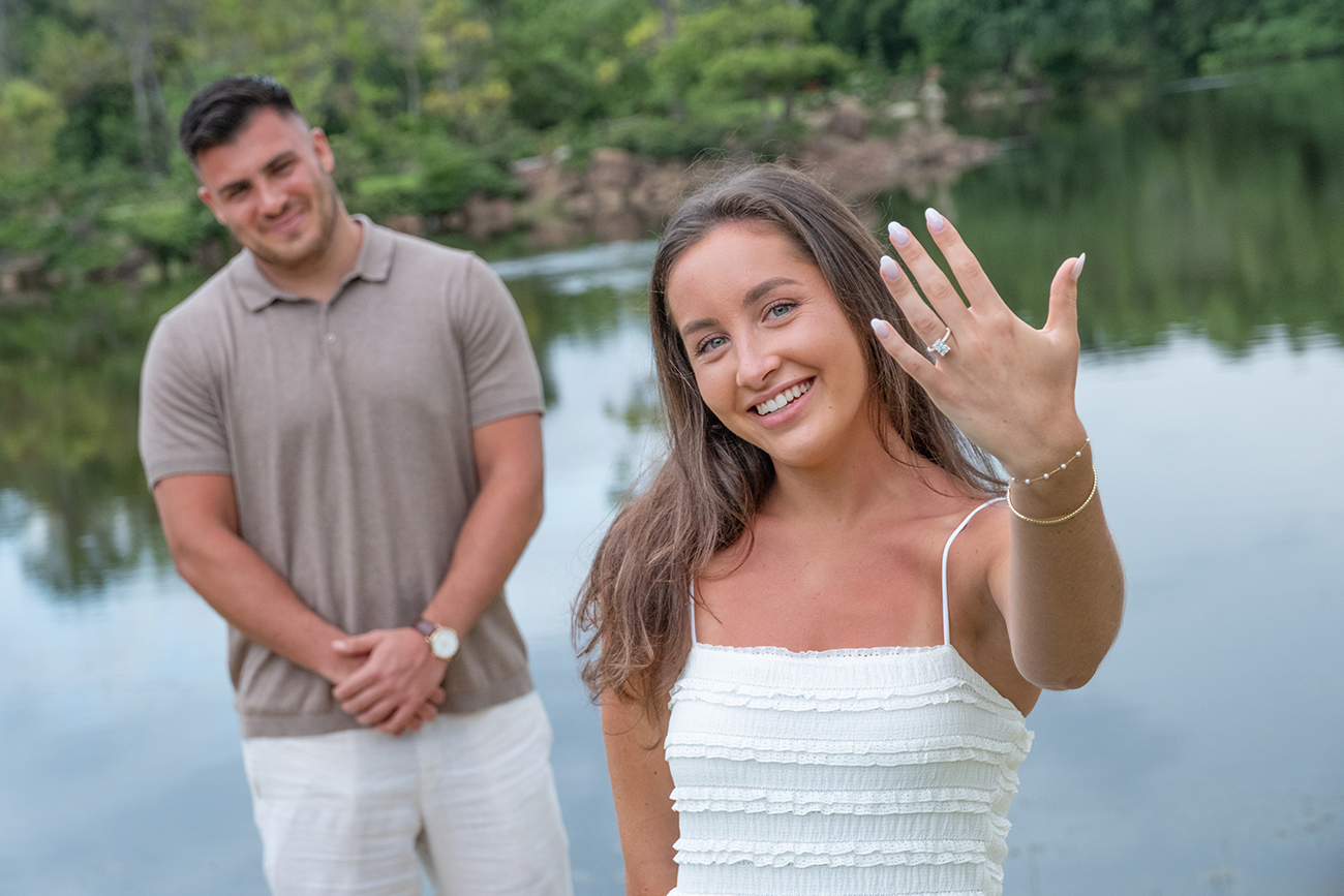 A newly engaged woman beams with joy as she shows off her engagement ring beside a peaceful lake at the Morikami Museum and Japanese Gardens in Delray Beach, Florida, while her fiancé smiles proudly in the background.
