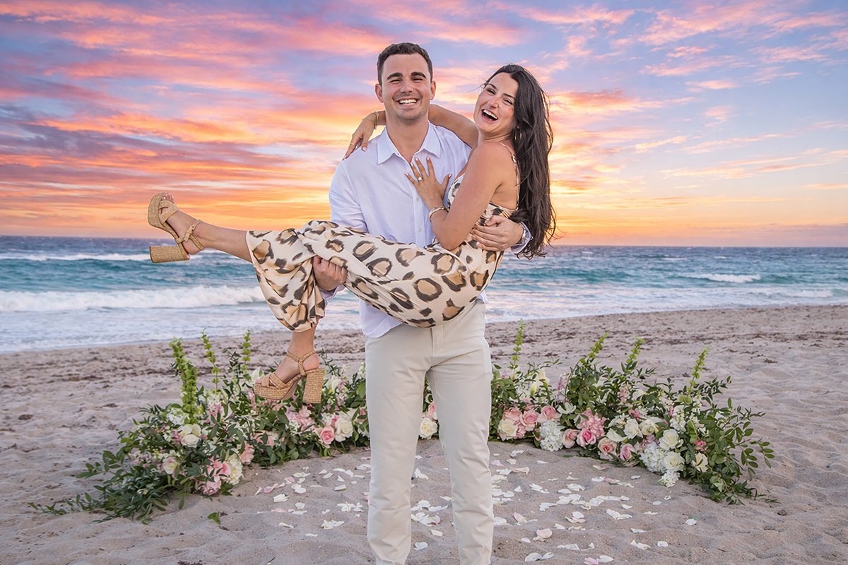 Couple celebrating after a beach proposal in Boca Raton at sunset, with the groom holding his fiancée in his arms in front of ocean waves and floral arrangements.