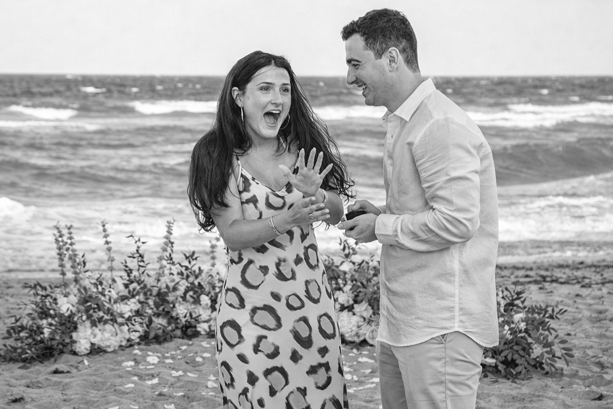 Black-and-white photograph of a marriage proposal on a beach in Boca Raton, Florida, showing a woman reacting with joyful surprise, her mouth open and hand raised as she looks at her engagement ring, while her fiancé smiles beside her holding the ring box. Ocean waves and floral arrangements frame the romantic scene.