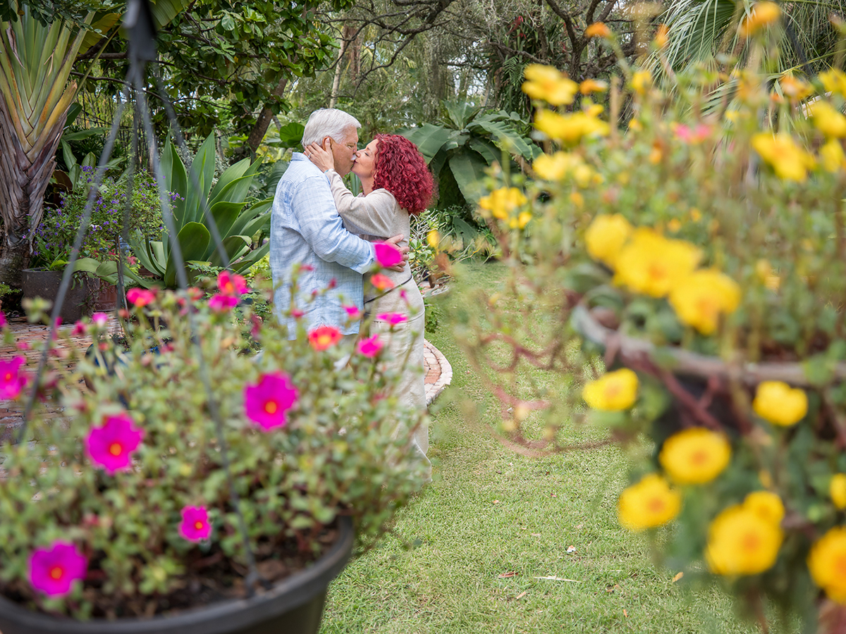 A mature, elegant couple sharing a gentle kiss in a lush Coral Gables home garden, framed by vibrant flowers and tropical greenery, evoking timeless romance and luxury at home.