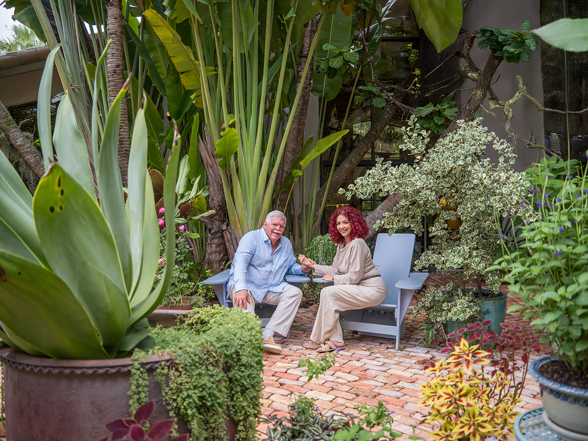 Premium backyard portrait of a mature South Florida couple seated in a lush tropical garden with tall palms, vibrant plants, and brick pathway, captured by a professional wedding and lifestyle photographer in natural light.