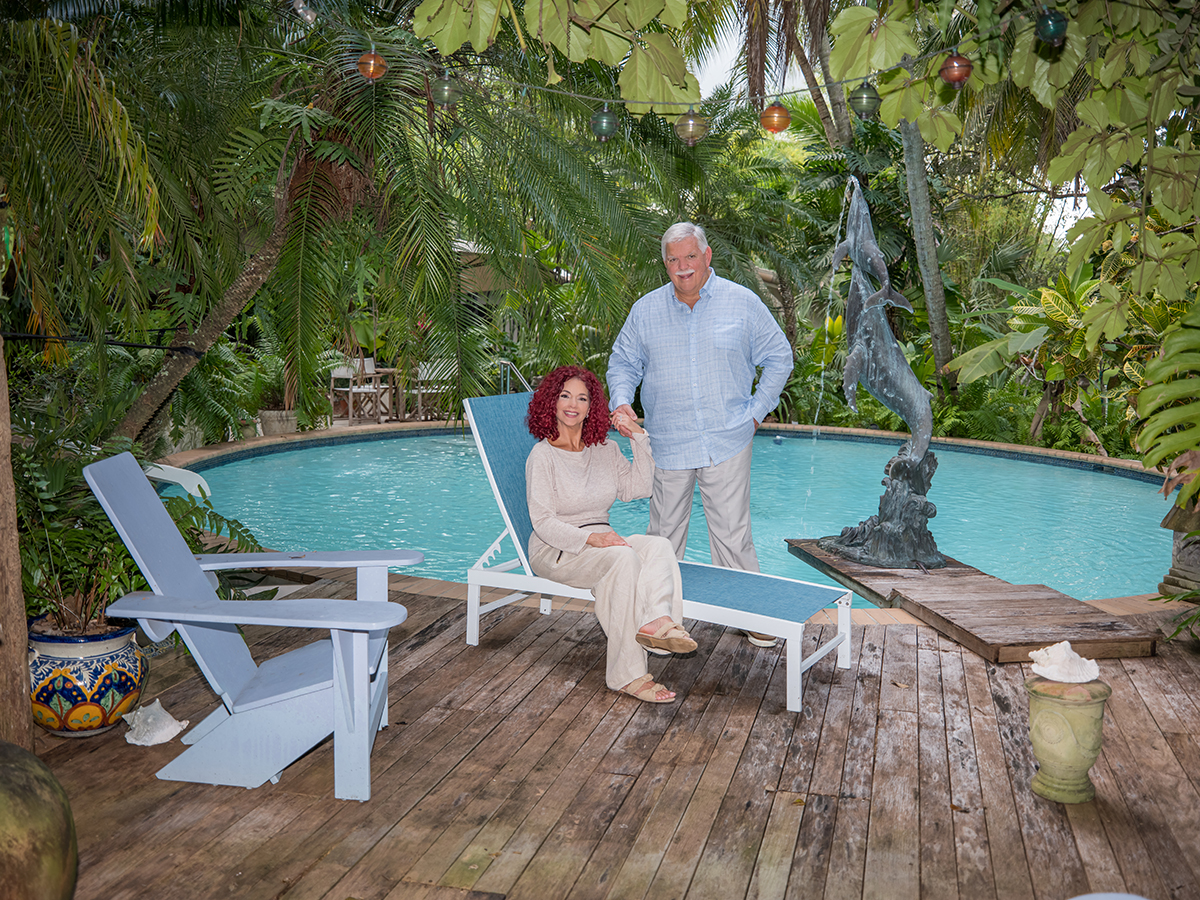 Mature couple holding hands by a resort-style backyard pool in South Florida, surrounded by lush tropical landscaping, outdoor lounge chairs, and decorative garden sculptures, photographed with soft natural light and a high-end lifestyle portrait aesthetic.