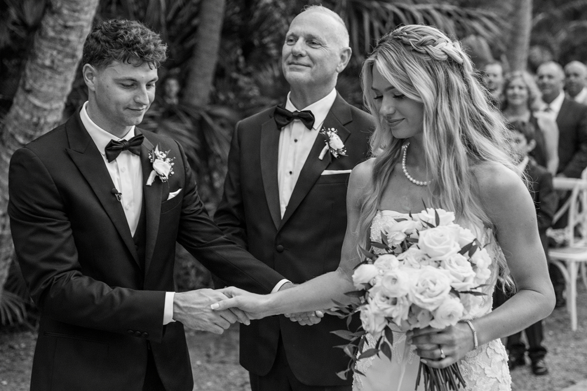 Black and white Bonnet House wedding ceremony photo of groom taking bride’s hand while her father stands proudly behind in Fort Lauderdale garden setting.