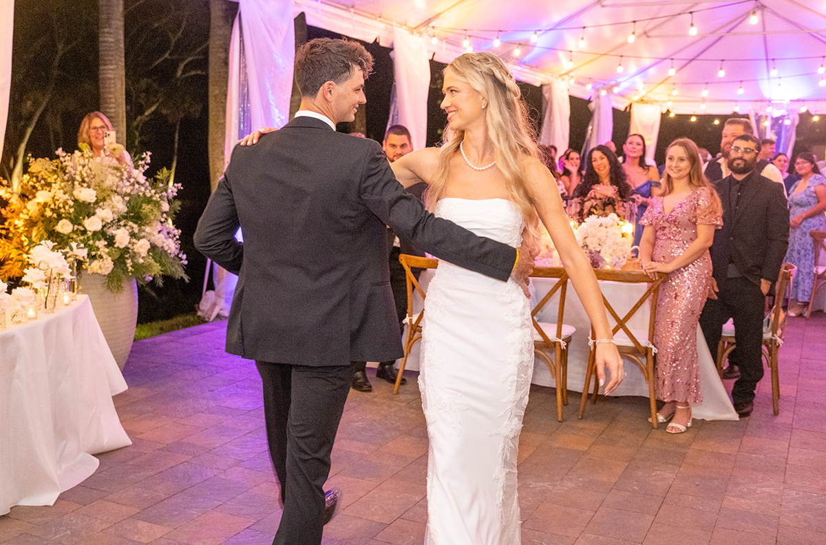 Bride and groom sharing their first dance at The Bonnet House in Fort Lauderdale under café lights with guests watching and candlelit floral décor surrounding them.