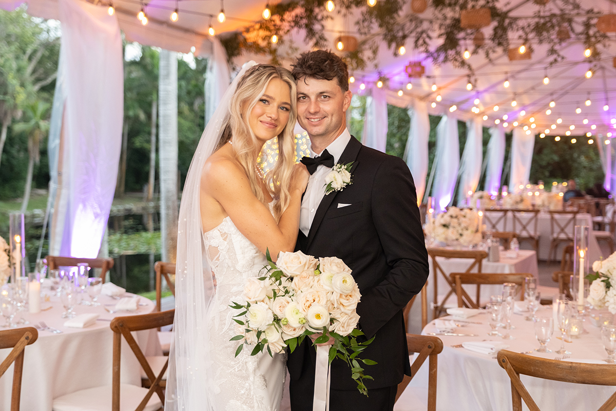 Bride and groom standing together inside a tented wedding reception at The Bonnet House in Fort Lauderdale with café lights, blush table linens, white floral centerpieces, and waterfront backdrop.