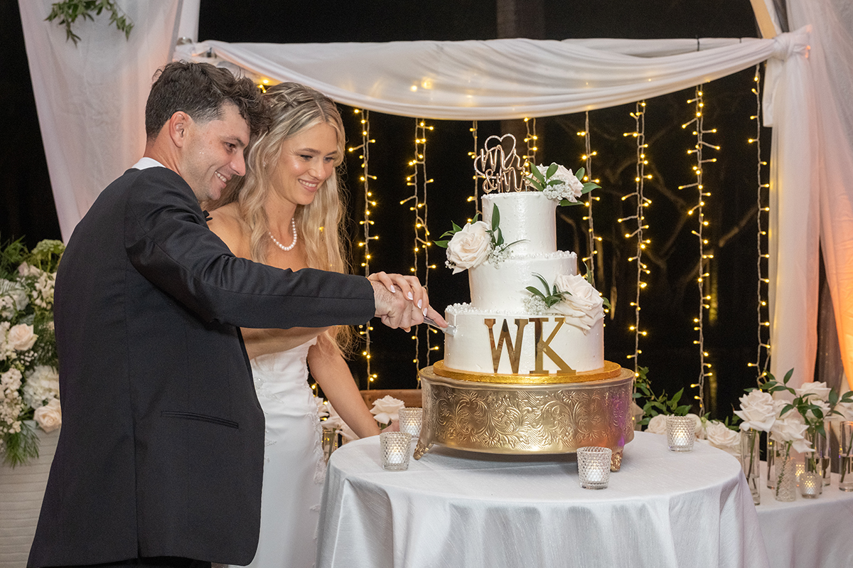 Bride and groom cutting a three-tier white wedding cake under café lights at the end of their Fort Lauderdale wedding reception.