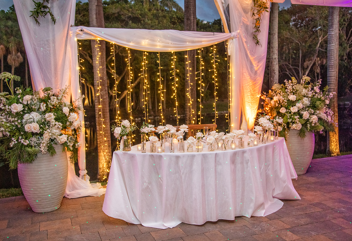 Wedding head table at The Bonnet House in Fort Lauderdale with white roses, tall floral arrangements, candlelight, and cascading string lights against a waterfront backdrop at sunset.