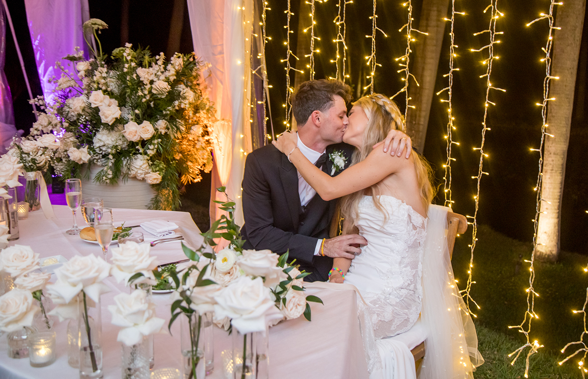 Bride and groom kissing at their head table during a nighttime wedding reception at The Bonnet House in Fort Lauderdale, surrounded by white roses and cascading café lights.