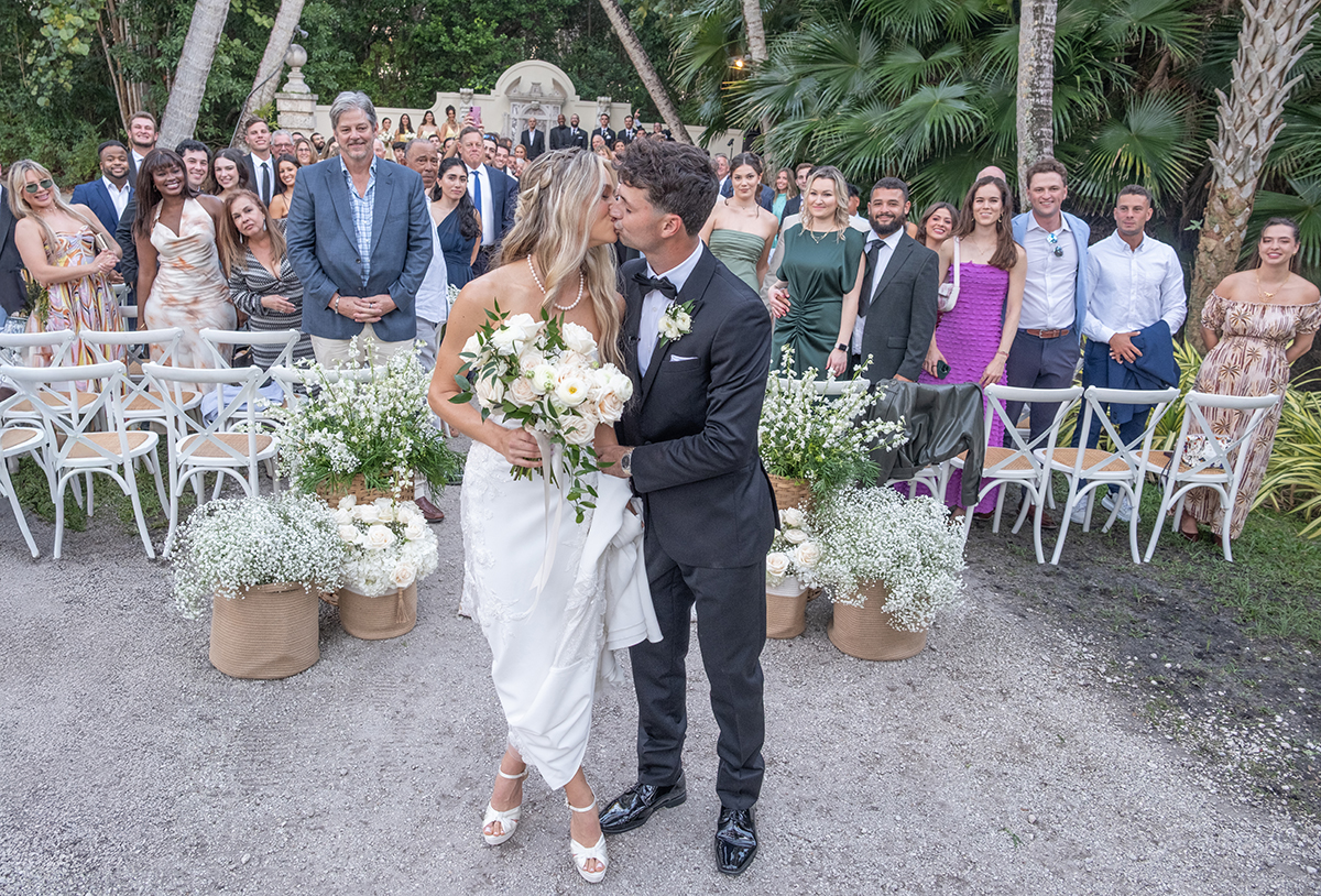 Bride and groom kissing at the end of their Bonnet House wedding ceremony in Fort Lauderdale surrounded by cheering guests and tropical greenery.