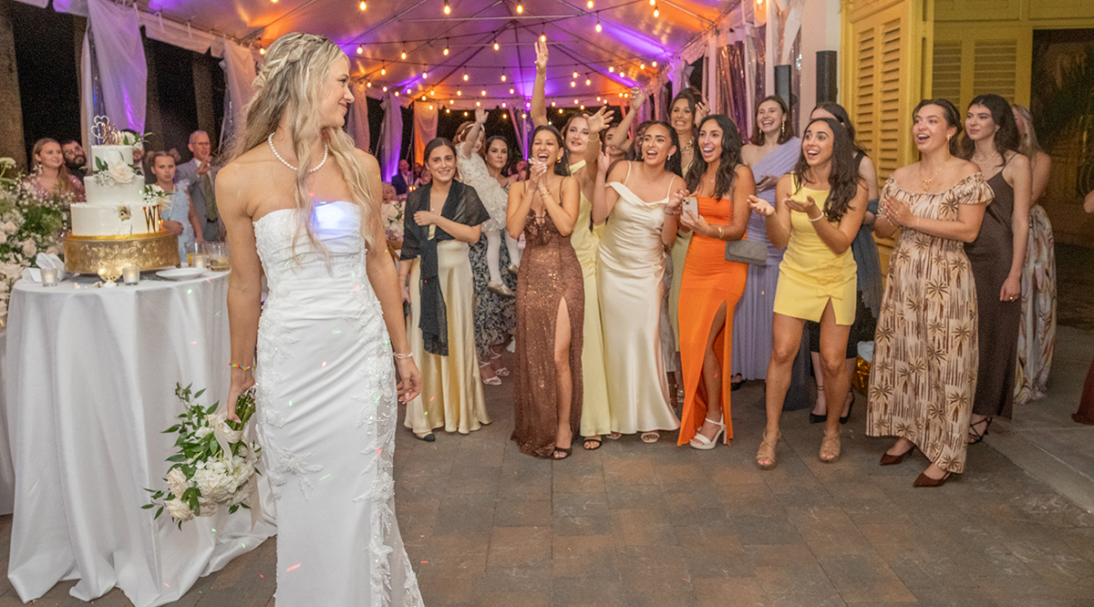 Bride looking over her shoulder before the bouquet toss at The Bonnet House in Fort Lauderdale while a group of women wait excitedly behind her under café lights.