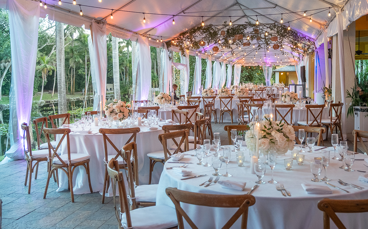 Elegant wedding reception tent at Bonnet House in Fort Lauderdale overlooking the lake, with white linens, blush floral centerpieces, wooden cross-back chairs, draped fabric, and glowing string lights.