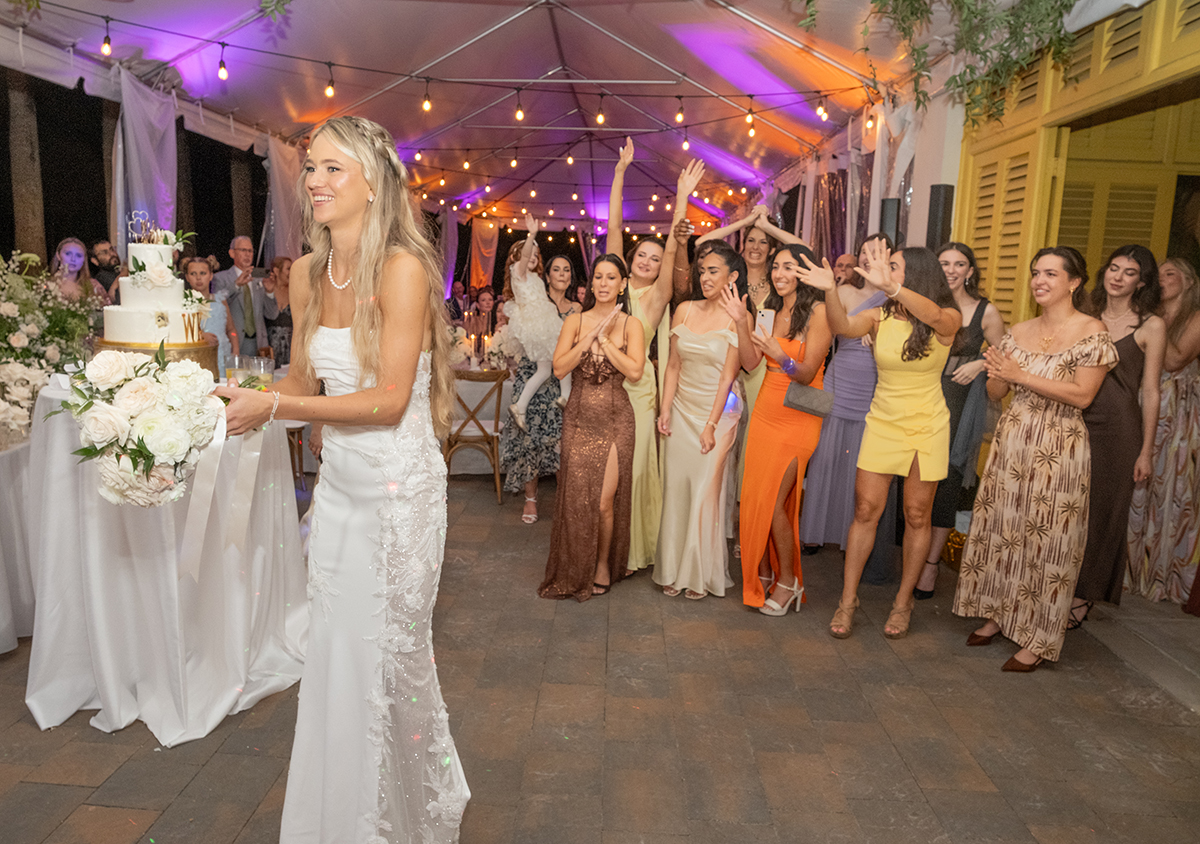 Bride holding bouquet and smiling before the bouquet toss at The Bonnet House in Fort Lauderdale while a group of women gather with hands raised behind her under café lights.