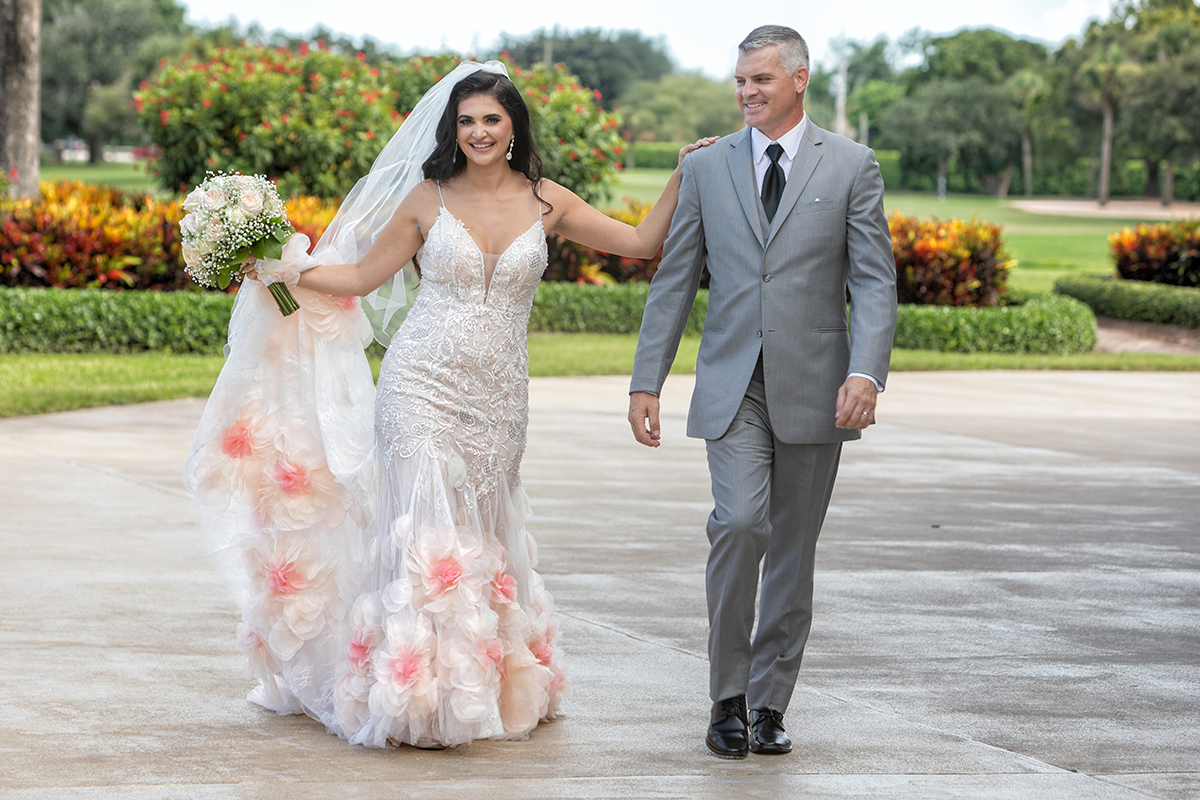 A joyful bride and groom walking together outdoors at The Club at Boca Pointe, highlighting the bride's unique mermaid-style gown with intricate lace and pink floral appliques.