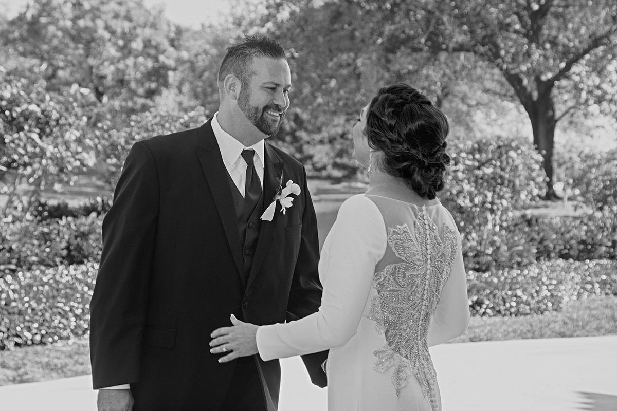 Black-and-white photo of a groom smiling at his bride during their first look at the Club of Boca Pointe, highlighting his expression and the intricate details of her gown.