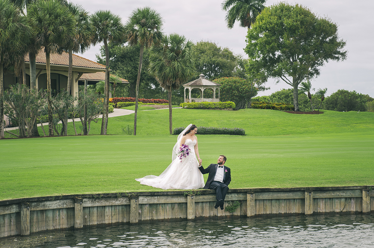Newlywed couple holding hands by a serene lake at The Club at Boca Pointe, surrounded by lush greenery and a gazebo in the background.
