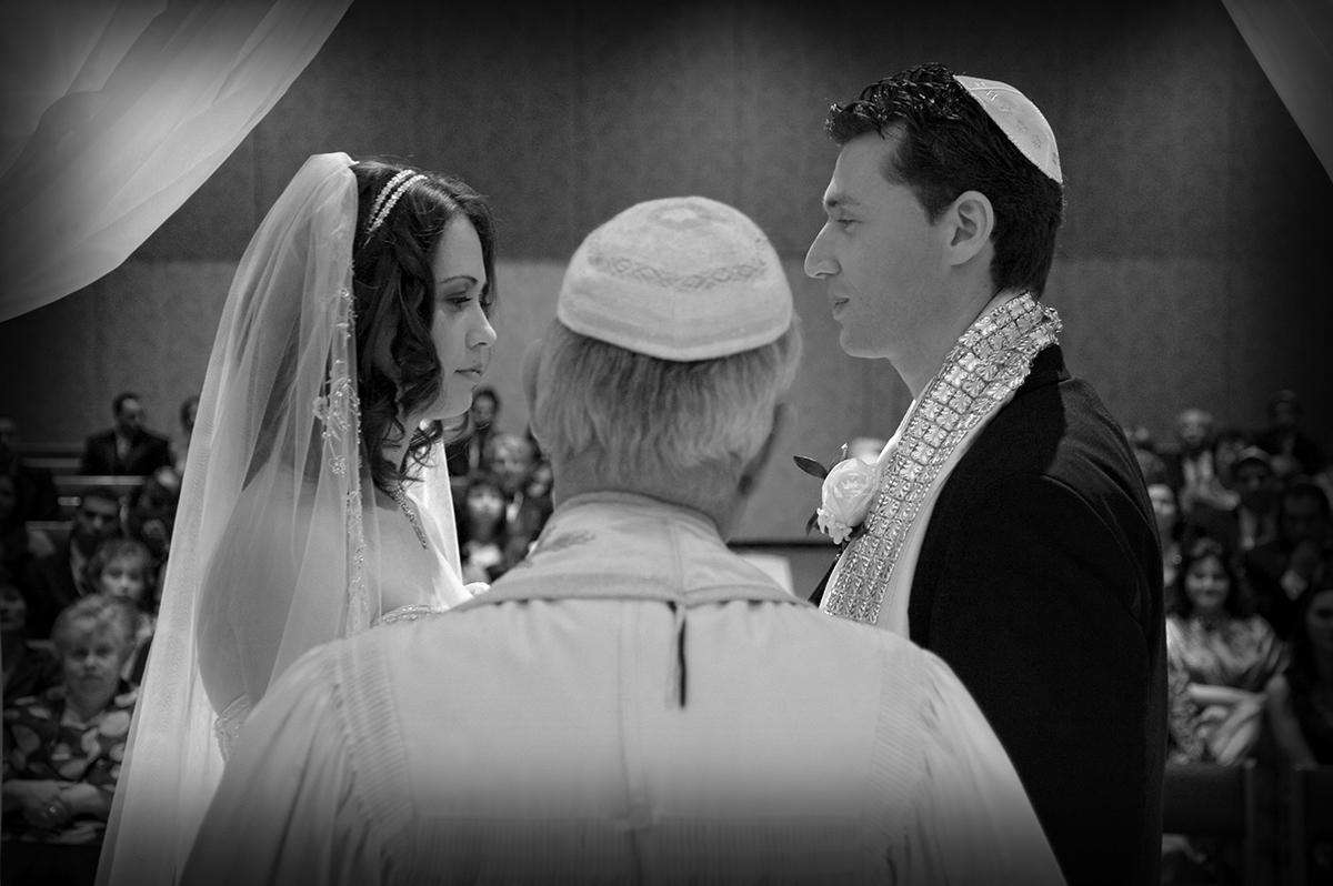Black and white photo of a Jewish wedding ceremony showing bride and groom facing each other under the chuppah with the rabbi in the foreground, captured using ambient light.