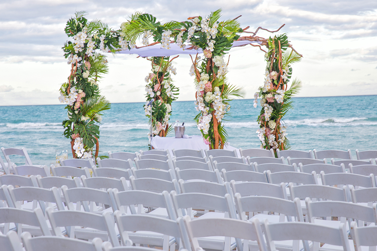 Elegant beach chuppah at Fontainebleau Hotel in Miami set against the ocean, decorated with tropical greenery and white flowers for a Jewish wedding ceremony
