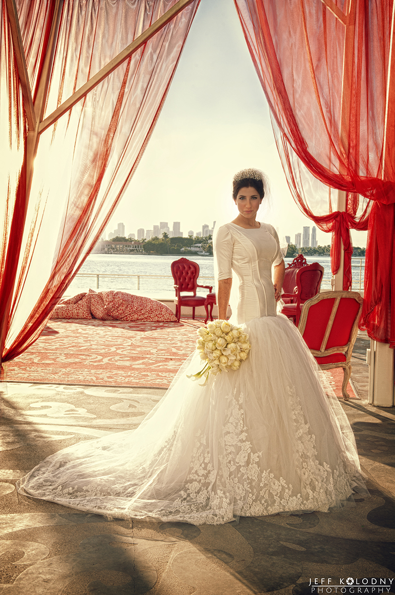 Bride in elegant lace wedding gown holding bouquet under red draped canopy with waterfront city skyline in background