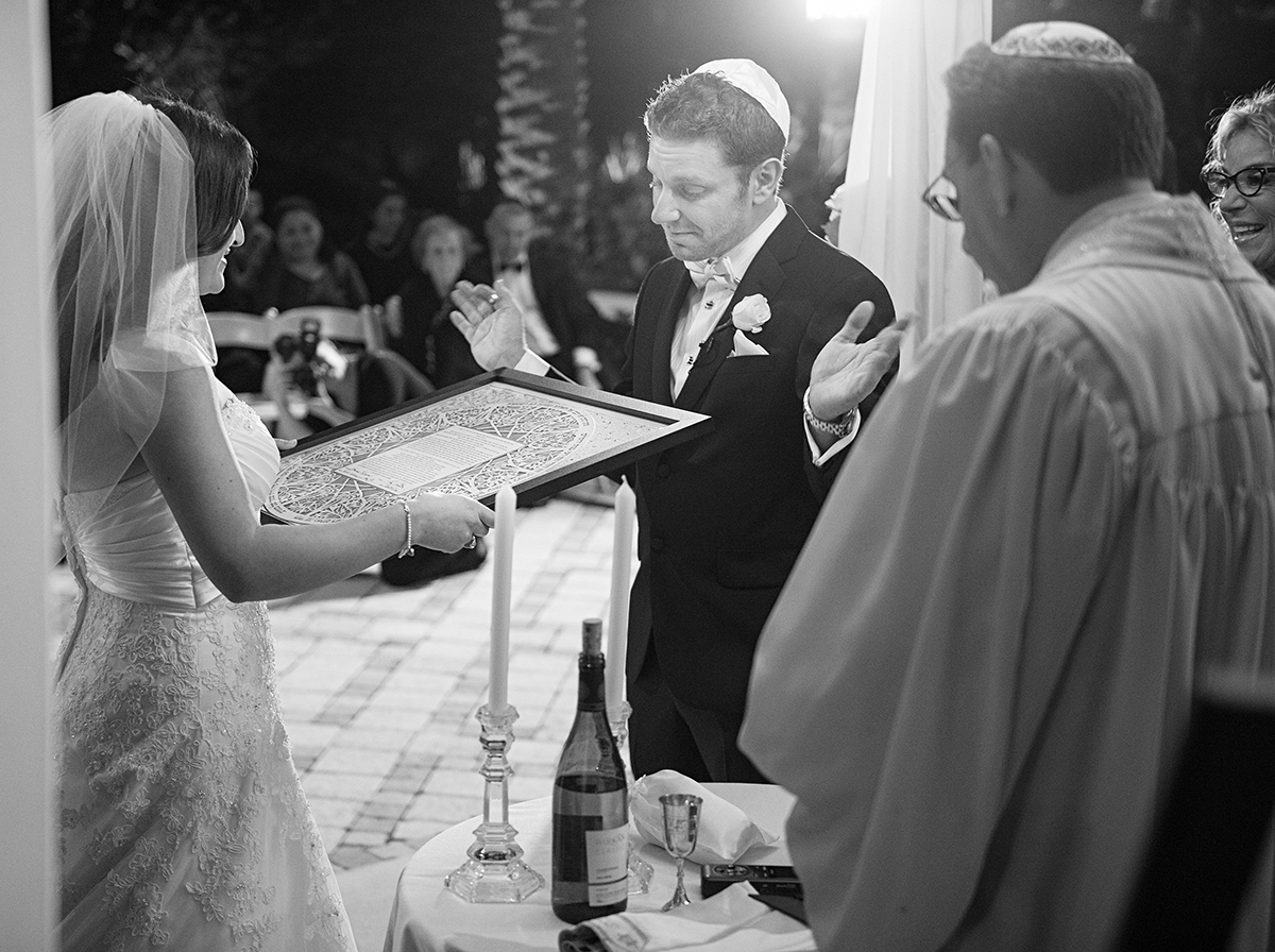Bride and groom holding ketubah during Jewish wedding ceremony under chuppah with rabbi and candles black and white image