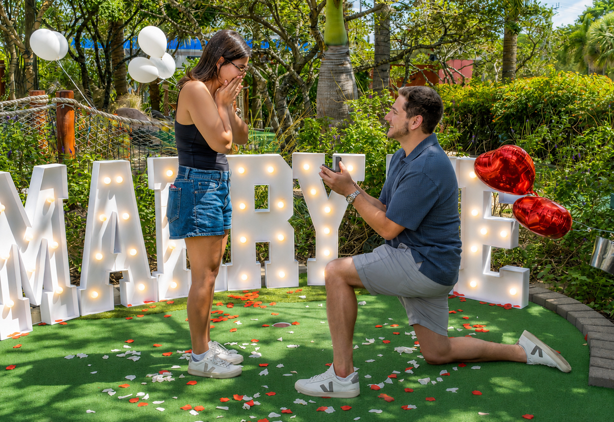 Jacob proposes to Lauren during a surprise marriage proposal at Putt'n Around Delray Beach as she reacts with emotion while standing in front of illuminated "Marry Me" letters surrounded by rose petals and balloons.