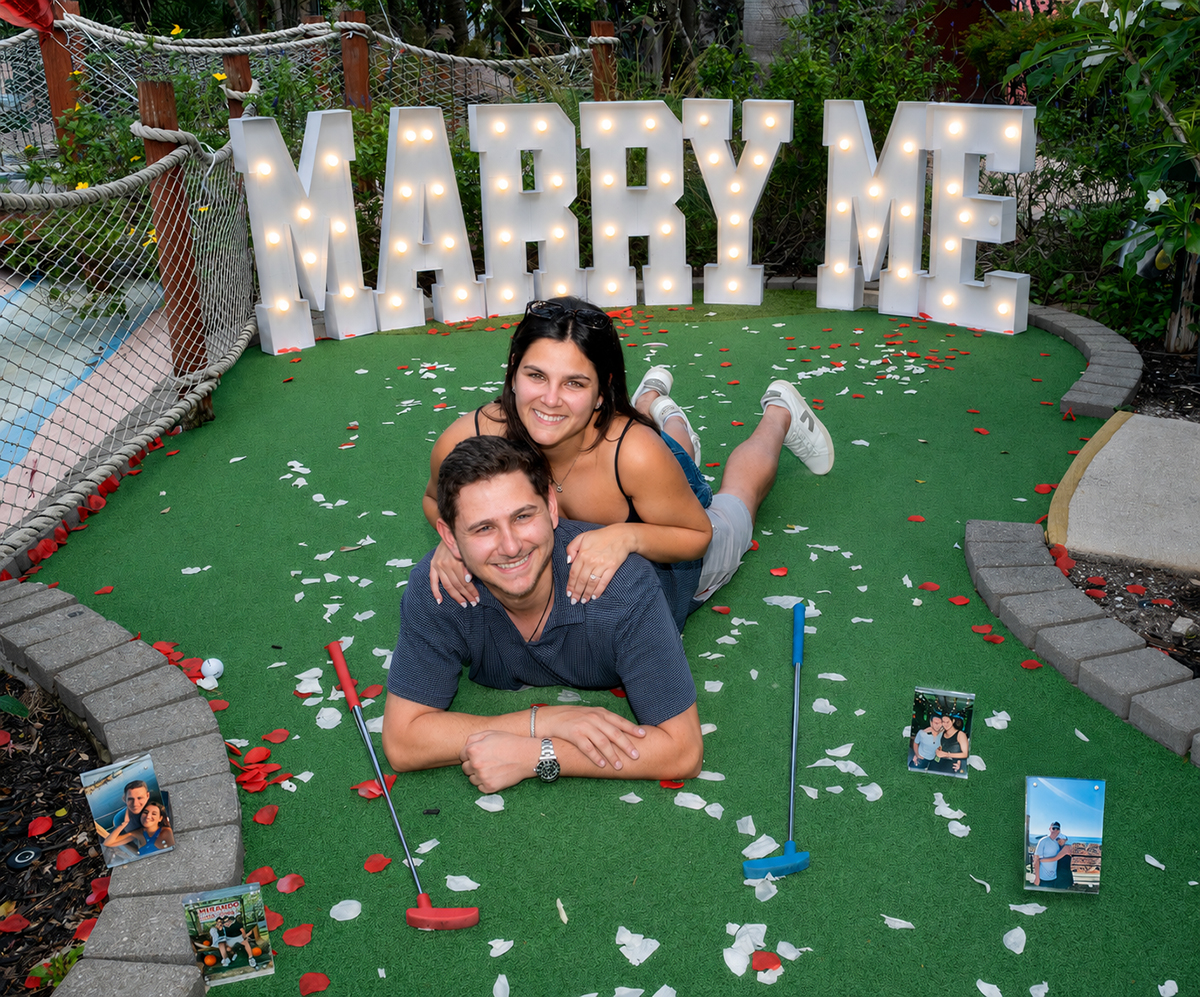 Jacob and Lauren pose together on a miniature golf green at Putt'n Around Delray Beach following their surprise marriage proposal, surrounded by rose petals, framed photographs, and illuminated "Marry Me" letters.