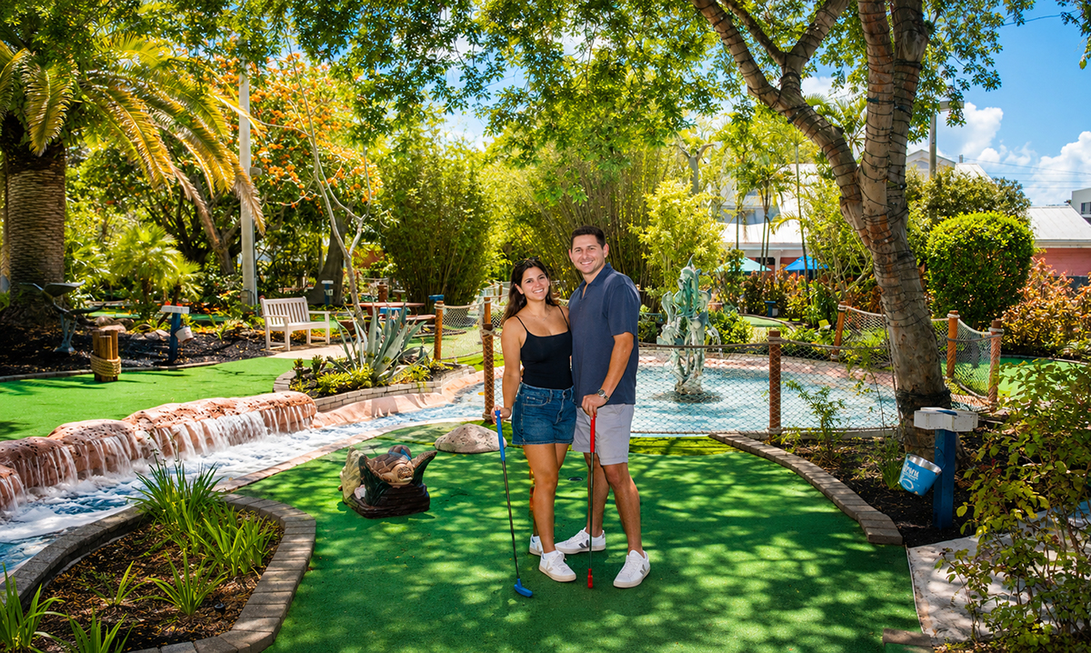 Jacob and Lauren posing together on a tropical miniature golf course at Putt'n Around Delray Beach after their surprise marriage proposal, surrounded by lush landscaping, waterfalls, and palm trees.