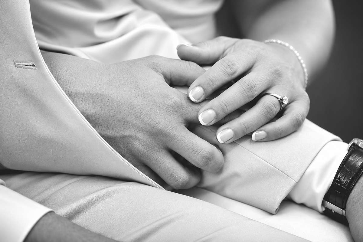 Close-up black-and-white photograph of Catarina's hand resting on David's arm during their Catholic wedding ceremony, highlighting her engagement ring and wedding day details.