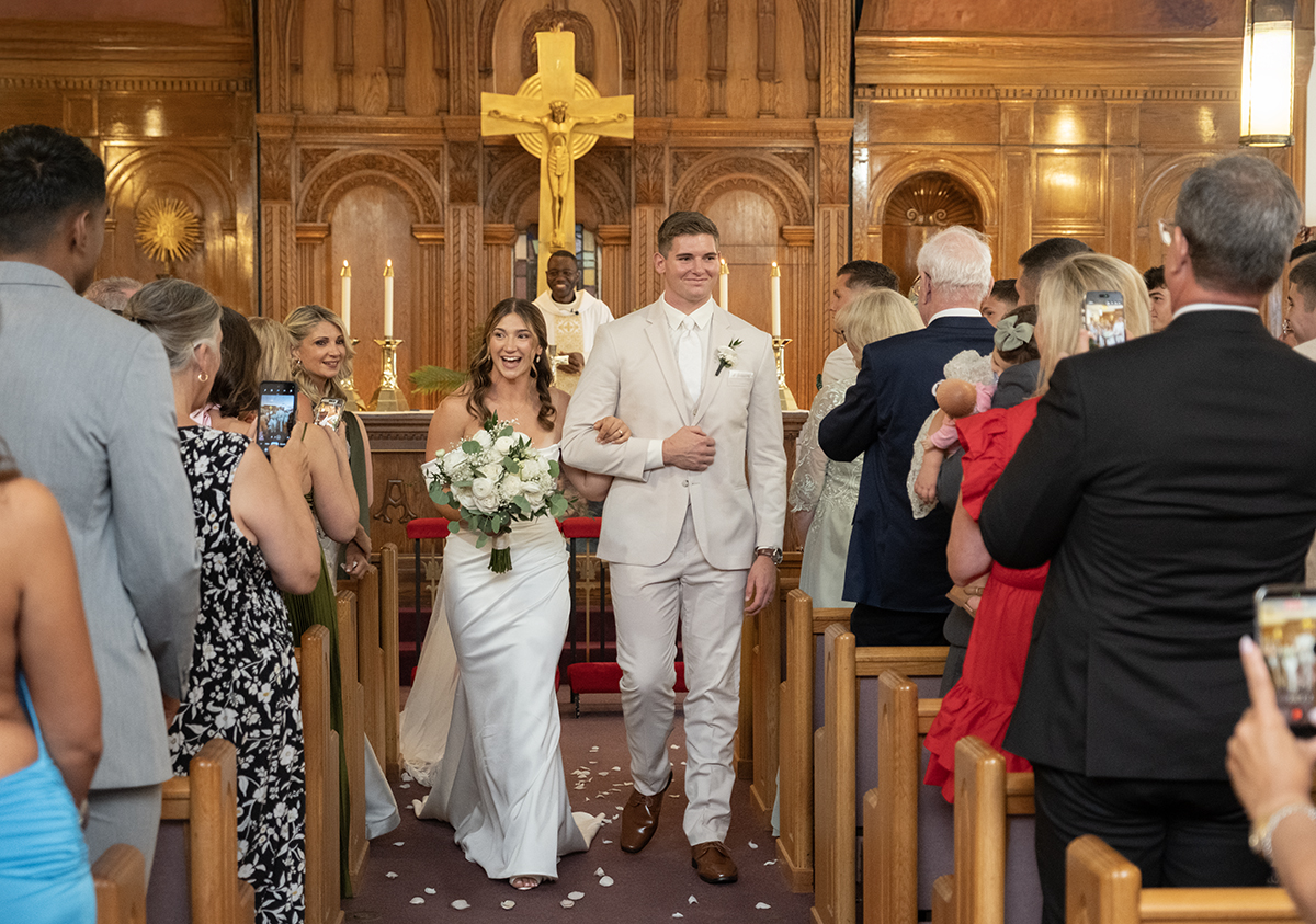 Newly married couple Catarina and David walk down the aisle after their Catholic wedding ceremony at Our Lady of Mercy Church in South Florida, greeting family and friends as husband and wife.