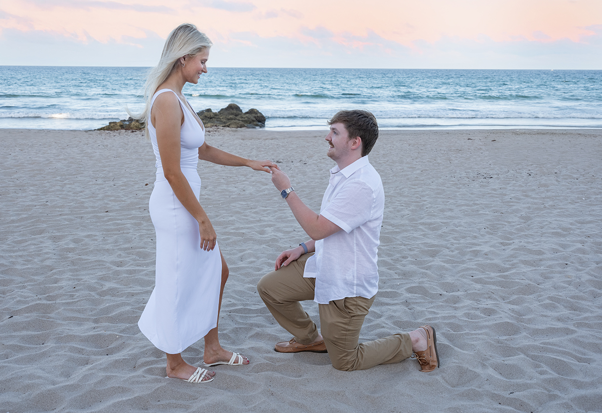 Josh on one knee proposing to Rachael on a Boca Raton beach with the ocean behind them
