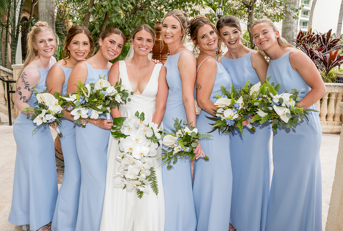 Bride surrounded by her bridesmaids in light blue dresses holding white, blue and green floral bouquets.